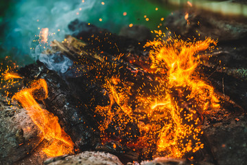 Vivid smoldered firewoods burned in fire close-up. Atmospheric background with orange flame of campfire and blue smoke. Warm full frame image of bonfire with glowing embers in air. Bright sparks bokeh