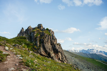 Beautiful mountain landscape with footpath and great sharp rock on hillside. Awesome alpine scenery with pointy crag on background of snowy mountains under blue sky. Trail near big pointed stone.