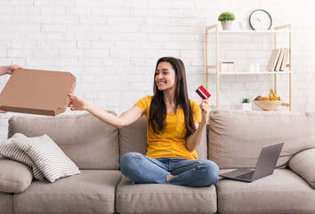 Online delivery. Young woman with credit card and laptop computer getting her ordered pizza from courier indoors