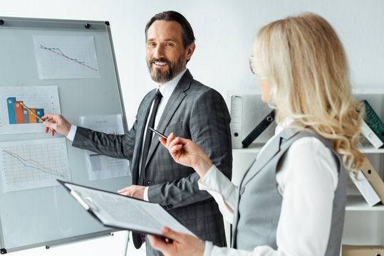 Selective focus of smiling businessman pointing on charts on whiteboard near businesswoman holding clipboard in office