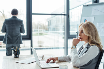 Selective focus of pensive businesswoman using laptop while businessman standing near window in office