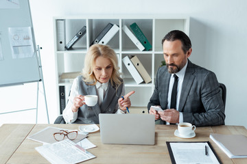 Businesswoman with cup of coffee pointing with finger at businessman with smartphone near laptop and documents on table