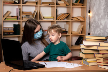 young mother in a medical mask with a small child sit at a desk with books and a laptop. quarantined distance learning.