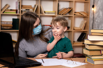 Fototapeta na wymiar mom in a medical mask with a little boy in her arms at a table with a laptop. family leisure. bookcase on the background. online learning