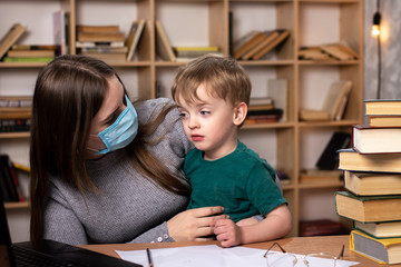 young mother in a medical mask holds a baby in her arms. sitting at a desk with books and a laptop. quarantine at home.
