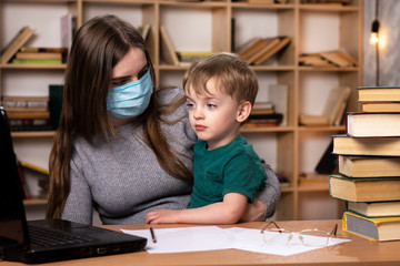 mother in a medical mask sits with her three-year-old son in her arms in an apartment. bookcase on the background. laptop and books on the table. self-isolation, quarantine.