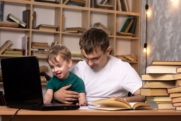 a young man in glasses holds a child in his arms. sitting in the library with a laptop. many books lie nearby. leisure dad and son