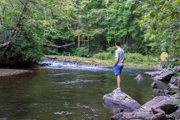 Young man in gray t-shirt and blu shorts is standing on big rock and looking on beautiful landscape with mountain river and green forest