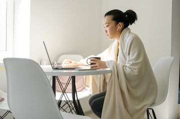 Asian woman wearing blanket. Young Asian woman working from home while sitting at a table. Asian girl using laptop.