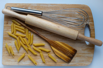Top view of uncooked penne spaghetti with wooden rolling pin, wooden spatula and metal steel wire whisk on wooden cutting board