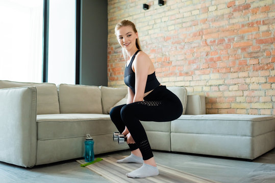 happy sportswoman doing squat exercise with dumbbells near sports bottle in living room