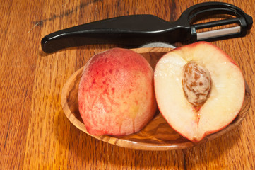 Top view, close distance of a freshly picked peach, cut in half to view pit, in a rare wood bowl, and a peeling utensils on a wood cutting board