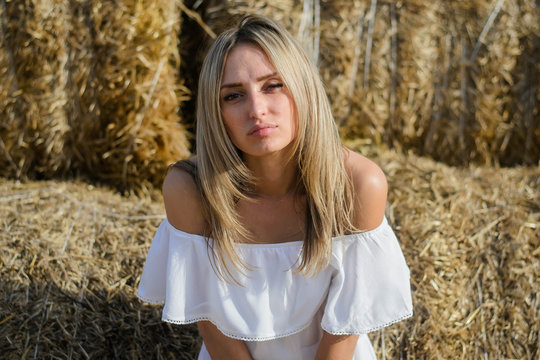 Provincial Young Woman Of Blonde, Of Eastern European Descent In A White Dress Posing Against A Background Of Haystacks. A Young Girl From The Village, A Haystack, A Farmer Girl, National Economy.