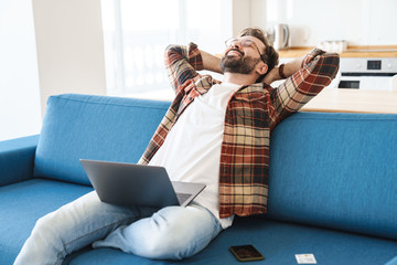 Portrait of young happy man smiling and using laptop on sofa at home