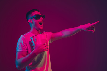 Pointing, holding popcorn. Caucasian man's portrait isolated on pink-purple studio background in neon light. Male model with devices. Concept of human emotions, facial expression, sales, ad. Copyspace