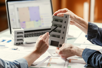 Two architects holding and discussing model of a house and architectural plans in a studio with blueprints and laptop on a table.