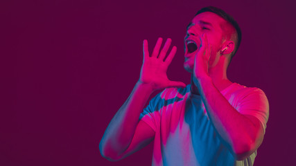 Naklejka na ściany i meble Calling, shouting. Caucasian man's portrait isolated on pink-purple studio background in neon light. Beautiful male model with earphones. Concept of human emotions, facial expression, sales, ad.
