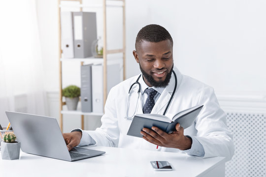 Busy african doctor working with laptop onlinr, checking his notebook