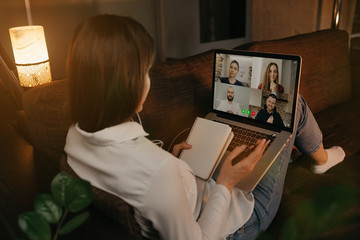 Back view of a woman lying at home on a sofa talking with her colleagues in a video call on a laptop. Businesswoman doing notes in a notebook during a video conference. A team having an online meeting