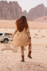 An elegant girl in cowboy boots and a short dress stands on a stone on one leg. Her dress was pouted by the wind. Back view. In the background a desert landscape