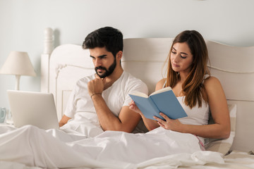 Young woman reading a book in bed while her boyfriend using laptop