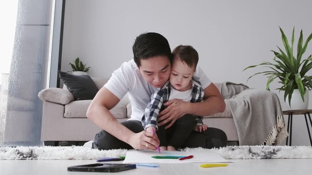 The parent is painting with his toddler child. The father is holding son’s hand and they are drawing together. The family is at home on quarantine.