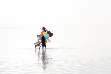 Girl playing guitar while sitting on the chair. Sunset, Sea reflection and colorful balloons 