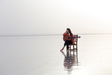Girl playing guitar while sitting on the chair. Sunset, Sea reflection and colorful balloons 