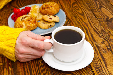 Female hands in yellow sweater holding cup of coffee and cookies on brown old wooden table.