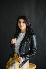 portrait of a young dark-haired girl on a dark background sitting on a bar stool with dark leather jacket and skirt. Photography of a young beautiful girl in the Studio