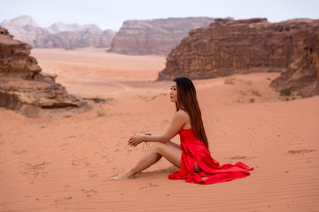 Fototapeta na wymiar A cute Asian girl with long hair in a red long dress sits on the sand and looks into the distance. In the background the desert and mountains