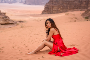 Fototapeta na wymiar a cute girl of Asian appearance in a red long dress sits on the sand in the desert and looks at the camera