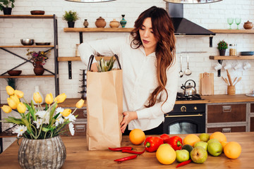 A young woman came back from a supermarket. There are many fruits and vegetables: apples, red peppers, hot peppers, oranges, pears in her bag. She is keeping pineapple in hands. Kitchen at home.