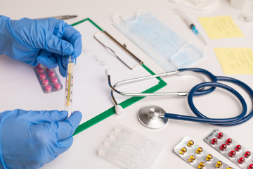 Doctor in the workplace. The doctor has a thermometer in his hands to measure the temperature. On the table phonendoscope, tablets, injection ampoules, syringes, copybook