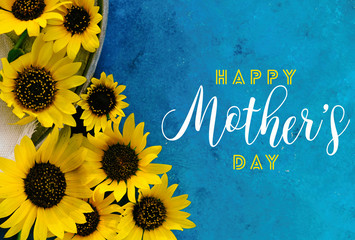 Happy Mothers Day background graphic with flat lay of sunflowers on blue texture.