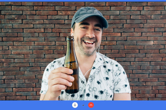 smiling man chatting in a video call using earphones - Happy and cool person drinking a beer with friends using technology during lockdown isolation