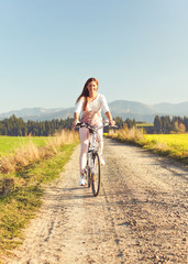 Fototapeta premium Young woman in summer light clothes rides bicycle on dusty road towards camera, afternoon sun shines to fields and forest in background