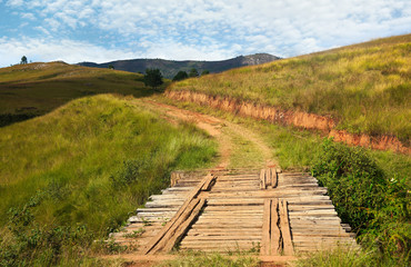 Fototapeta na wymiar Bridge made of wooden bars on country road, grass growing on hills both sides - typical view when driving 4wd off road vehicle near Andringitra park, Madagascar
