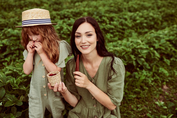 Beautiful young mother with her daughter having fun on a green field with strawberries