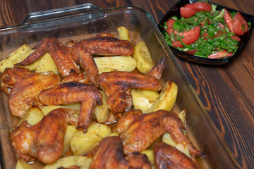 Baked chicken wings with fried potatoes served on the wooden table with a plate of tomato salad