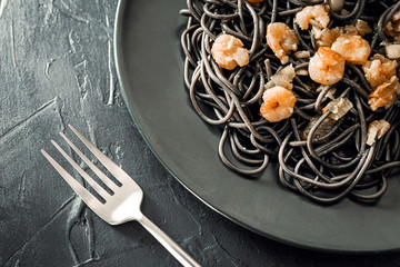 Flat lay of elegant black plate of pasta with sepia ink and prawns next to a silver fork with black textured background