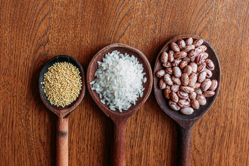 White rice, pinto beans and proso millet seeds on wooden kitchen spoons.