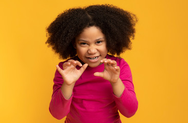 Little black girl imitating tiger, roaring and grimacing over yellow background