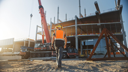 Back Shot of Worker Contractor Wearing Hard Hat and Safety Vests Walks on Industrial Building Construction Site. In the Background Crane, Skyscraper Concrete Formwork Frames 