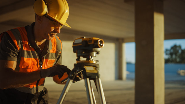 Inside of the Commercial / Industrial Building Construction Site: Professional Engineer Surveyor Takes Measures with Theodolite, Using Digital Tablet. In Background Skyscraper Formwork Frames Crane 