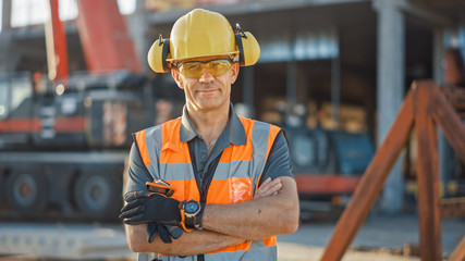 Portrait of Successful Builder / Worker / Contractor Wearing Hard Hat and Safety Vest Standing on a Commercial Building Construction Site, Crosses Arms Confidently. In the Background Crane Machinery