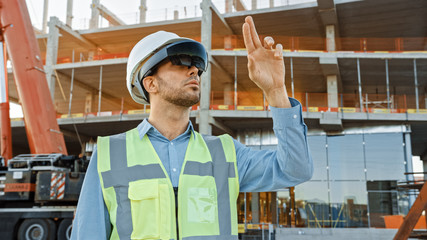 Futuristic Architectural Engineer Wearing Augmented Reality Headset and Using Gestures to Control Commercial Building Construction Site. In Background Skyscraper Formwork Frames and Industrial Crane