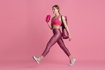 Fototapeta na wymiar Beautiful young female athlete with bag and water bottle in studio, monochrome pink portrait. Sportive fit caucasian model before training. Body building, healthy lifestyle, beauty and action concept.
