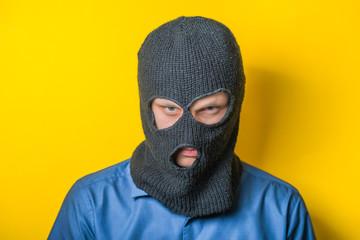 man close up thief in a mask and a blue shirt on a yellow background looks slyly to the camera. Mimicry. Gesture. photo Shoot/ evil criminal wearing balaclava - 341778374