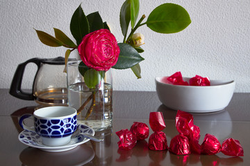 Recipiente con bombones de chocolate envueltos en papel rojo, una taza de café y una flor